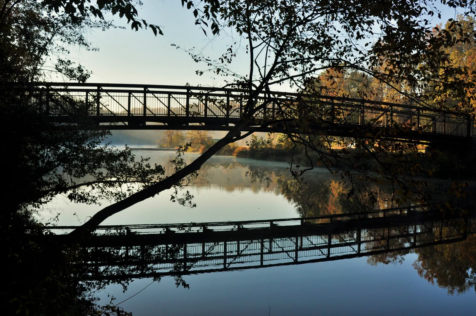 Pasarela peatonal sobre un río en calma al amanecer, reflejada en el agua entre árboles otoñales